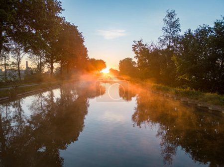 Experience the enchanting beauty of dawn as fiery fog blankets a tranquil canal, illuminated by the gentle rays of the rising sun. This captivating scene evokes a sense of serenity and wonder, as