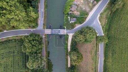 Photo for Experience the enchanting beauty of nature from above with this top-down aerial view. Roads converge toward a charming drawbridge that spans a serene canal meandering through lush forests and fertile - Royalty Free Image