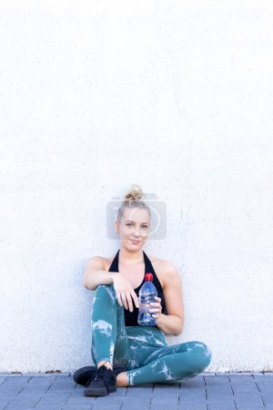 Foto de Una joven rubia feliz y deportiva milenaria se sienta en el suelo con una botella de agua después de hacer ejercicio, irradiando bienestar y un compromiso con un estilo de vida saludable mientras mira a la cámara - Imagen libre de derechos