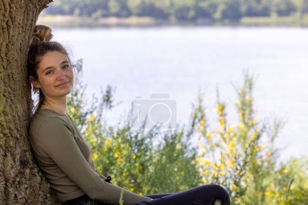 Téléchargez les photos : Une jeune femme brune trouve réconfort dans la forêt luxuriante, regardant un lac bleu tranquille, embrassant la sérénité d'une journée d'été. Méditation Serene Forest Lake. Photo de haute qualité - en image libre de droit