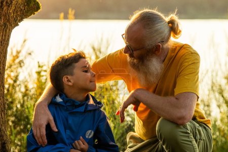 Ein herzerwärmender Moment, als ein liebevoller Großvater und sein kleiner Enkel bei Sonnenuntergang im Gras an einem Waldsee sitzen und eine besondere Verbundenheit in der ruhigen Schönheit der Natur teilen. Großvater und Enkel