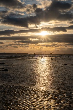 Téléchargez les photos : Un moment à couper le souffle capturé alors que le soleil doré se couche sur une plage française pittoresque, jetant des teintes chaudes sur le rivage, avec de petites vagues roulant doucement sous des nuages spectaculaires. Coucher de soleil doré à - en image libre de droit