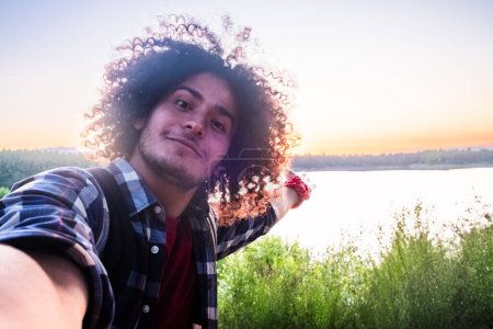 Téléchargez les photos : Un homme arabe joyeux avec de longs cheveux bouclés est sur un séjour de randonnée le long d'un sentier de lac de forêt tranquille. Avec la douce lueur du coucher de soleil coulant une teinte dorée, il capture un selfie, son sourire rayonnant - en image libre de droit