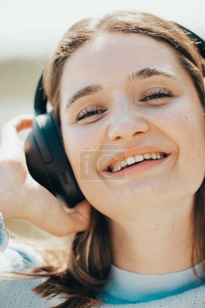 Photo for With the sea as her backdrop, a young woman in casual summer attire exudes excitement, her headphones a gateway to an auditory escape. The sunlight plays upon her features, highlighting a carefree - Royalty Free Image