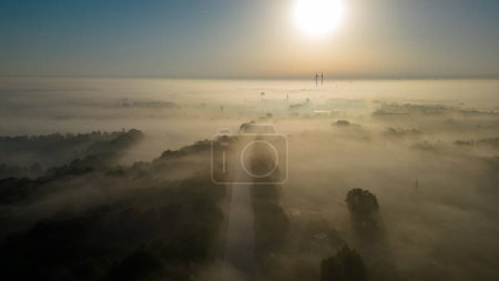Photo for This serene image captures the tranquil beauty of a countryside scene at dawn. The sun hangs low in the hazy sky, casting a warm glow that permeates the mist. Below, the fog enshrouds the landscape - Royalty Free Image