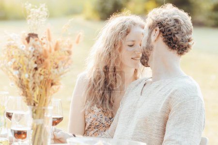 The photograph captures a tender moment between a couple, leaning in close for a moment of connection at an outdoor table. Their faces nearly touching, they share a gaze that speaks to a deep