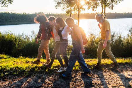 Téléchargez les photos : Cette image animée représente un groupe d'amis marchant de façon ludique le long d'un chemin au bord du lac au coucher du soleil. Leur mouvement transmet un sentiment d'énergie et de plaisir insouciant. Les torches chaudes du soleil à travers - en image libre de droit