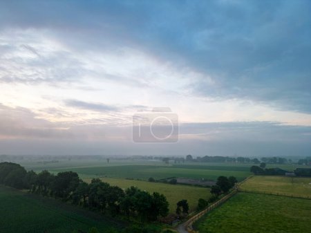 Photo for This image presents a tranquil early morning scene over the countryside, with the first light of dawn breaking across the horizon and casting a gentle glow over the landscape. The patchwork of fields - Royalty Free Image