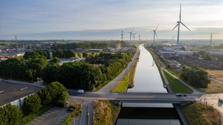 The image provides an aerial perspective of a canal running through an urban landscape, with wind turbines rising in the background. The turbines stand as sentinels of sustainable energy, their sleek