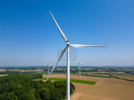 Foto de La fotografía muestra una majestuosa turbina de viento que domina el horizonte rural, sus cuchillas alcanzando el cielo azul claro. De pie en medio de una mezcla de campos cultivados y vegetación natural, el - Imagen libre de derechos