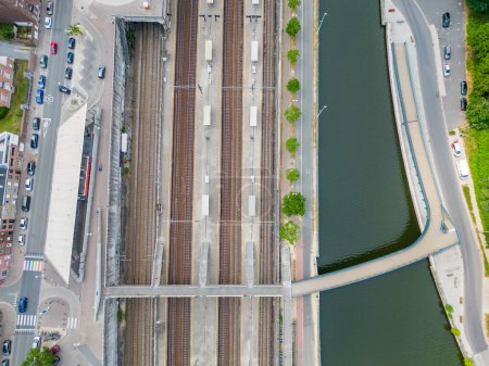 La imagen presenta una vista de arriba hacia abajo de una estación de tren adyacente a un río curvo, que ilustra la confluencia de diferentes modos de tránsito. Las vías del tren corren paralelas, marcadas por plataformas