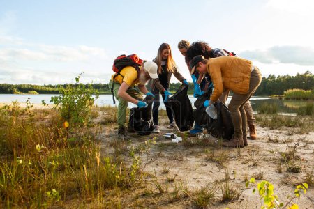 Esta imagen captura a un grupo de individuos que participan activamente en una limpieza junto al lago. Vestidos con atuendo casual al aire libre y armados con bolsas de basura y guantes, se doblan, recogiendo basura