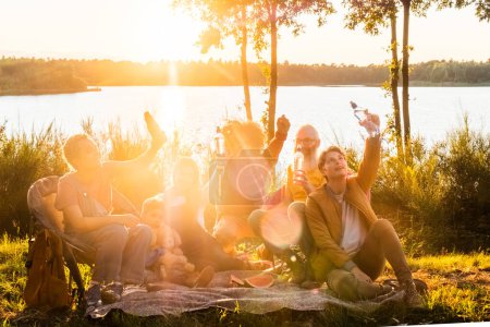 La imagen está impregnada de la cálida luz dorada del sol poniente, capturando a un grupo de amigos disfrutando de una vibrante reunión junto al lago. Brazos levantados y sonrisas anchas, se deleitan en la alegría de la