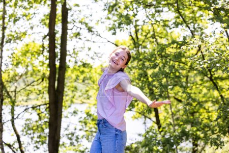 Photo for The image features a young woman with a playful pose, her arms outstretched, dancing or twirling in a forest setting. Her carefree spirit is infectious, and her smile is as bright as the dappled - Royalty Free Image