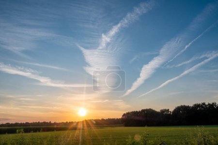 Foto de La imagen captura una majestuosa puesta de sol sobre un campo verde, donde el sol besa el horizonte, enviando rayos de luz a través del cielo. Las nubes dramáticas están grabadas en la extensión azul, formando - Imagen libre de derechos