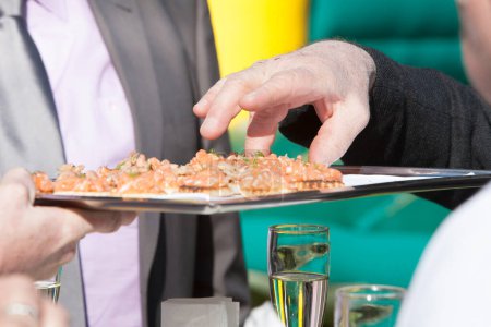 Photo for The image captures a moment at a catered event, with a server presenting a tray of smoked salmon canapes. An attendee is in the process of picking one of the delicately prepared appetizers - Royalty Free Image