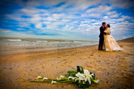 Foto de La imagen representa un momento conmovedor entre una novia y un novio, abrazándose en una playa de arena con el vasto y dinámico océano detrás de ellos. El cielo es dramático, con nubes que se separan para permitir rayos de luz - Imagen libre de derechos