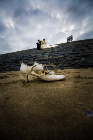 Téléchargez les photos : Cette image dramatique offre une perspective créative sur une scène de mariage à la plage, avec les chaussures de mariée au premier plan, abandonnées sur le sable alors que le couple échange des vœux au loin. L'angle bas - en image libre de droit