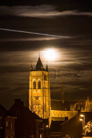 Foto de La iglesia de Sint-Lenaerts en Bélgica se levanta majestuosamente contra el lienzo nocturno, su estructura histórica iluminada por el resplandor inquietante de una luna llena. Los rayos de nube se deslizan por la cara lunar - Imagen libre de derechos
