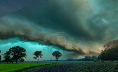A formidable shelf cloud dominates the horizon in this image, signaling the imminent arrival of a powerful storm over a serene farmland. The dark, undulating base of the cloud contrasts with the