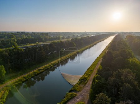 Téléchargez les photos : Cette image aérienne tranquille capture le calme d'un canal à l'aube, le soleil matinal projetant une lueur chaude à la surface des eaux. La voie navigable, flanquée de sentiers et d'arbres, sert de - en image libre de droit