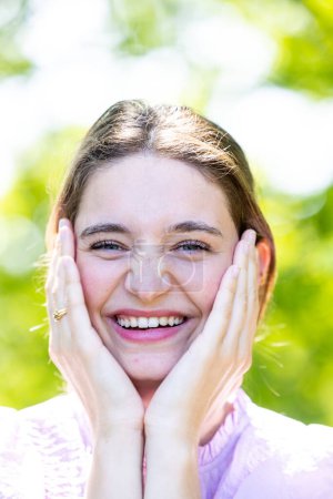 Photo for This image captures a young woman experiencing a moment of pure joy outdoors. She is holding her cheeks with her hands, and her expression is one of genuine delight and happiness. The woman is smile - Royalty Free Image