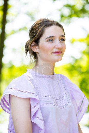 This portrait captures a young woman in a moment of contemplation. The natural light filters through the leaves, highlighting her soft features and the delicate details of her lavender blouse. Her