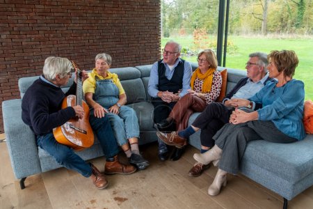 Dieses herzerwärmende Bild fängt eine Gruppe Senioren ein, die sich in einem gemütlichen Wohnzimmer versammelt und einen intimen musikalischen Moment genießt. Ein Herr wiegt eine Gitarre und erzählt vielleicht ein Lied oder eine Geschichte aus dem