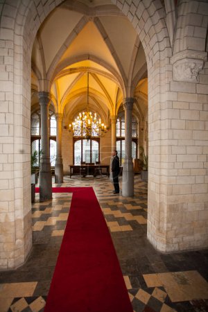 Foto de Esta fotografía captura la grandeza de un pasillo de estilo medieval, con arcos góticos y bóvedas acanaladas. Una lujosa alfombra roja se extiende por el centro, que conduce a una habitación cálidamente iluminada al final. - Imagen libre de derechos