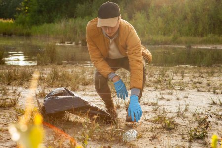 Photo for The photo captures a single volunteer, a young Caucasian man, engaged in a conservation effort, cleaning up a lakeside area. Hes bending down to pick up trash, wearing blue gloves to protect his - Royalty Free Image