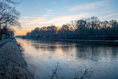 Photo for A tranquil early morning scene where a gentle sunrise graces the sky with pastel colors above an icy canal. The trees, devoid of leaves, stand as silent witnesses to the start of the day, their - Royalty Free Image