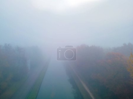 Foto de En esta fotografía, un canal atraviesa directamente el marco, flanqueado a ambos lados por árboles en pleno otoño. Los colores del follaje son apagados por una niebla densa y envolvente que suaviza la - Imagen libre de derechos