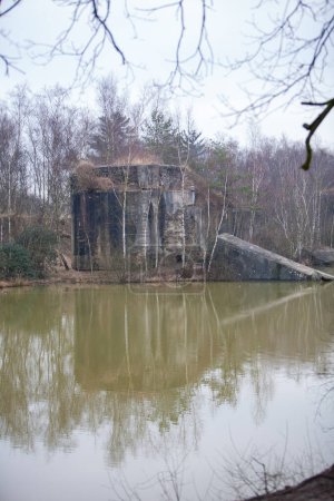 Téléchargez les photos : Cette image présente une vue sérénissime d'une structure abandonnée reflétée dans les eaux calmes d'un étang. Le bâtiment, enveloppé par les branches nues des arbres environnants, se dresse comme une relique de - en image libre de droit