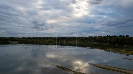 Photo for The image provides a sweeping view of a calm body of water reflecting a cloudy sky, with an autumnal forest in the background. The waters surface mirrors the grey clouds and patches of blue sky - Royalty Free Image