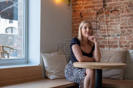 The image presents a young Caucasian woman with blonde hair sitting thoughtfully in a cozy corner of a stylish urban cafe. The setting combines modern and rustic elements, featuring a smooth wooden
