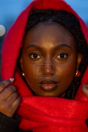 Téléchargez les photos : Ce portrait capture le regard intense et énigmatique d'une jeune femme africaine enveloppée dans une écharpe rouge vibrante. La photographie est prise pendant l'heure bleue, qui jette une teinte sereine, contrastant - en image libre de droit