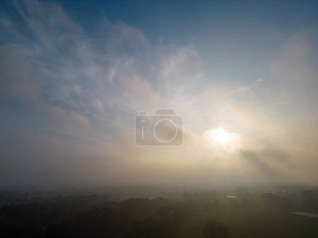 The image captures a stunning sunrise as the central focus, with the suns rays piercing through a dramatic sky filled with a tapestry of clouds. The mist that lingers over the rural landscape below