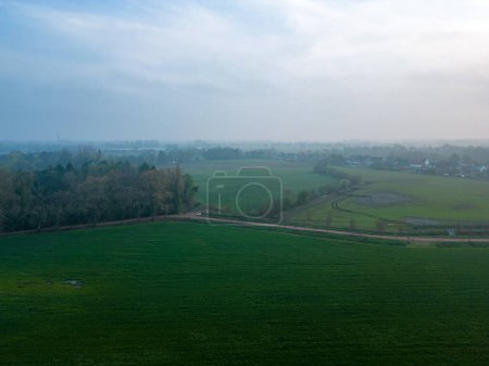 Photo for The image captures the serene essence of a misty morning over a sprawling farmland. The soft light of dawn diffuses through the haze, casting a gentle glow over the green fields. The distant horizon - Royalty Free Image