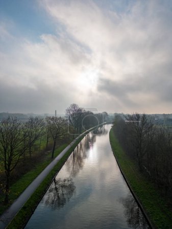 This image captures a tranquil morning scene over a river, taken from an elevated perspective. The soft sunlight diffuses through the hazy sky, reflecting gently on the waters surface. Flanking the
