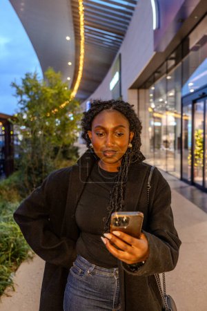 Photo for In the soft glow of twilight, a young Black woman is the focal point amidst the architectural lines of an urban shopping promenade. Her attire, a textured black overcoat atop a dark turtleneck - Royalty Free Image