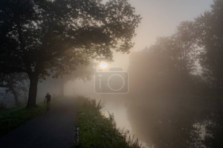 Téléchargez les photos : Un cycliste solitaire s'embarque dans un voyage le long d'un chemin riverain enveloppé de brouillard dans cette image évocatrice. Le soleil du matin, une sphère sourde, traverse le brouillard, projetant une douce lueur sur la scène - en image libre de droit