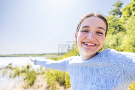 Foto de La imagen captura a una joven alegre en un suéter azul claro, extendiendo su brazo como si mostrara algo en la distancia en un lugar pintoresco junto al lago. Su brillante sonrisa y gesto abierto transmiten una - Imagen libre de derechos