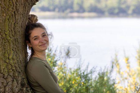 Photo for A young brunette millennial woman finds serenity in natures embrace. She sits by a forest lake, leaning against a tree, enjoying the tranquility of the lush green surroundings on a summer day - Royalty Free Image