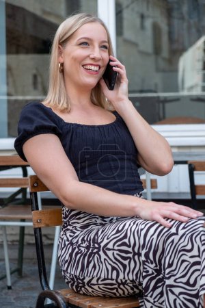 Dieses Bild zeigt eine fröhliche blonde Frau, die in einem Café im Freien lebhaft telefoniert. Ihr strahlendes Lächeln und das Funkeln in ihren Augen vermitteln das Glück des Austausches. Sie ist