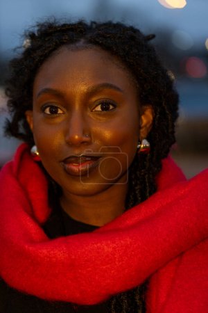 Foto de Este retrato captura a una joven y equilibrada mujer negra envuelta en una vibrante bufanda roja. La imagen emana calidez y sofisticación, resaltada por el suave efecto bokeh de las luces de las ciudades en el - Imagen libre de derechos