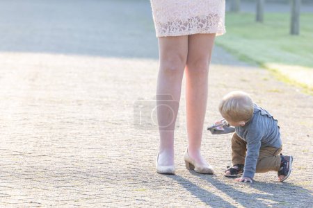 Photo for This image captures the essence of childhood curiosity as a toddler crouches down to explore the ground, with a young Caucasian woman standing close by, providing silent support. The morning sunlight - Royalty Free Image