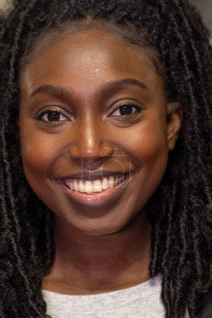 Ce portrait représente une jeune femme noire au sourire radieux et ouvert qui illumine le cadre. Ses cheveux sont coiffés dans des rebondissements naturels, encadrant magnifiquement son visage. Le scintillement dans ses yeux et le