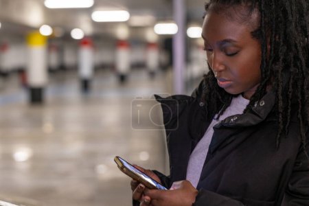 Una joven mujer está parada en una estación de metro aparentemente vacía, su atención completamente capturada por su teléfono inteligente. La suave iluminación de la estación arroja un brillo en su rostro, destacando su compromiso con