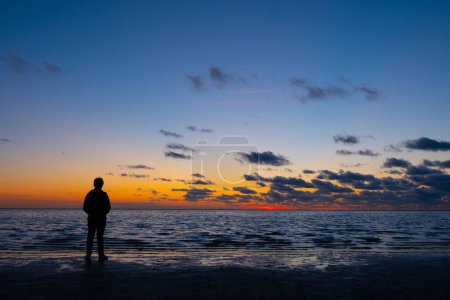 Téléchargez les photos : Cette image tranquille capture la silhouette d'un individu solitaire debout sur le rivage, plongé dans la sérénité du crépuscule. L'horizon est peint avec des teintes chaudes d'orange et de jaune, s'estompant - en image libre de droit