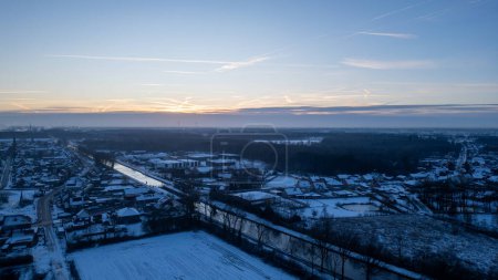 As dawn breaks, this aerial image captures a small town wrapped in the embrace of winter. The sunrise stretches across the horizon, spilling a soft light that gently graces the rooftops and frosted
